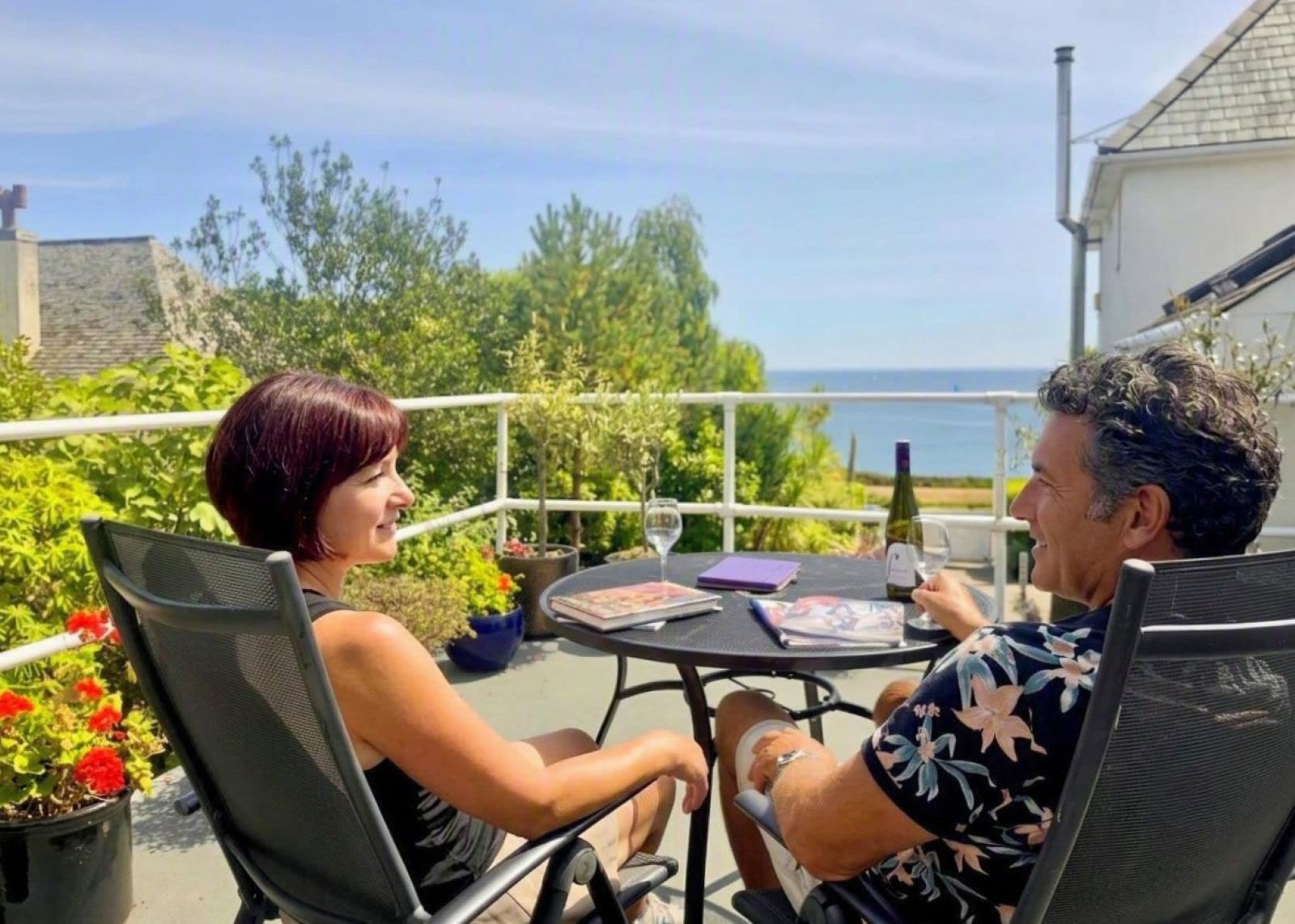 Two people sitting either side of a table on the private terrace at Blue Hills, with wine, and books, looking out towards Falmouth Bay